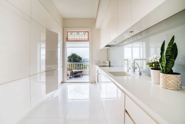 Bright, narrow contemporary kitchen with glossy white full-height cabinetry, white benchtops, integrated appliances, and a reflective pale floor. A long island or peninsula runs along the right side with a sink and mixer tap, while a glazed door at the rear opens to a balcony or outdoor area. Decorative stained-glass transom windows and indoor plants add character.