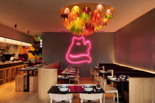 A warmly lit contemporary restaurant interior with an open kitchen and timber service counter on the left, rows of dining tables and chairs, curved timber partitions, dark wall panelling, and a bright pink neon cat-like wall sign. A large colourful sculptural pendant light dominates the ceiling, creating a playful focal point.