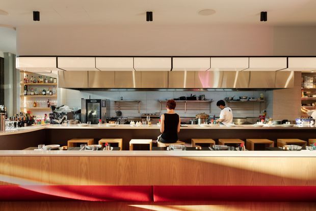 A contemporary commercial restaurant interior viewed from the dining area toward an open kitchen and service counter. The space features extensive warm timber cabinetry and panelling, a long bar-height counter with stools, stainless-steel cooking equipment and shelving, integrated lighting, and a prominent red upholstered banquette in the foreground.