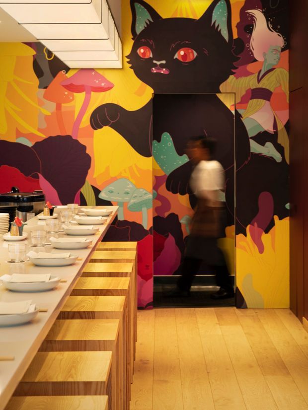 A vibrant restaurant interior with a long communal dining counter and stools, white tableware, warm timber flooring and cabinetry, and a large, colourful illustrated mural featuring a stylised black cat, mushrooms, foliage and figures. A dark reflective doorway or mirrored panel is integrated into the mural wall, with a blurred person passing through the space. The atmosphere is playful, bold and highly graphic.