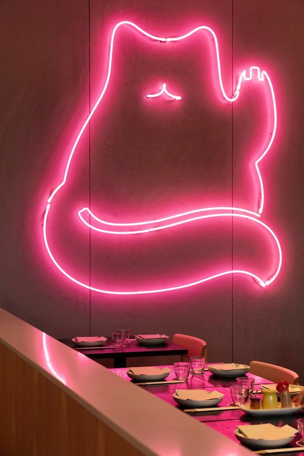 A dimly lit restaurant dining area featuring a large pink neon cat-shaped wall light above a long communal dining table. The table is set with plates, glasses, cutlery, and condiments, with warm wood panelling and pink ambient lighting creating a playful, dramatic atmosphere.