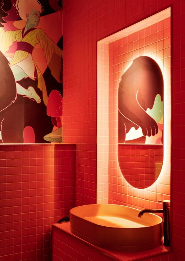 A dramatic, richly lit bathroom clad in small red tiles, featuring a sculptural oval vessel basin with dark tapware. A large illuminated recessed wall niche frames a bold abstract artwork, while additional graphic artwork appears on the adjacent wall. The space has a highly saturated red-orange palette and theatrical ambient lighting.