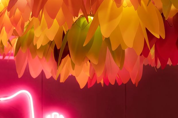 Close-up interior detail of a dramatic suspended installation made from layered, leaf-shaped translucent panels in vivid orange, yellow, red, and pink tones. The panels create a dense canopy overhead against a deep burgundy wall, with a partially visible glowing pink neon light at the lower edge. The image is highly saturated and atmospheric, with decorative feature lighting and an immersive, theatrical quality.