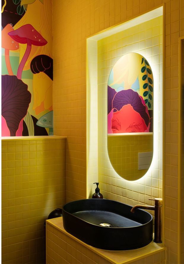 A compact, boldly designed bathroom with mustard-yellow square wall tiles throughout, a black vessel basin on a tiled vanity, and a brass-toned tap. An illuminated arched mirror is set into a tiled recess, reflecting vibrant botanical artwork with oversized colourful leaves and figures. The space has a highly saturated, playful and theatrical character.