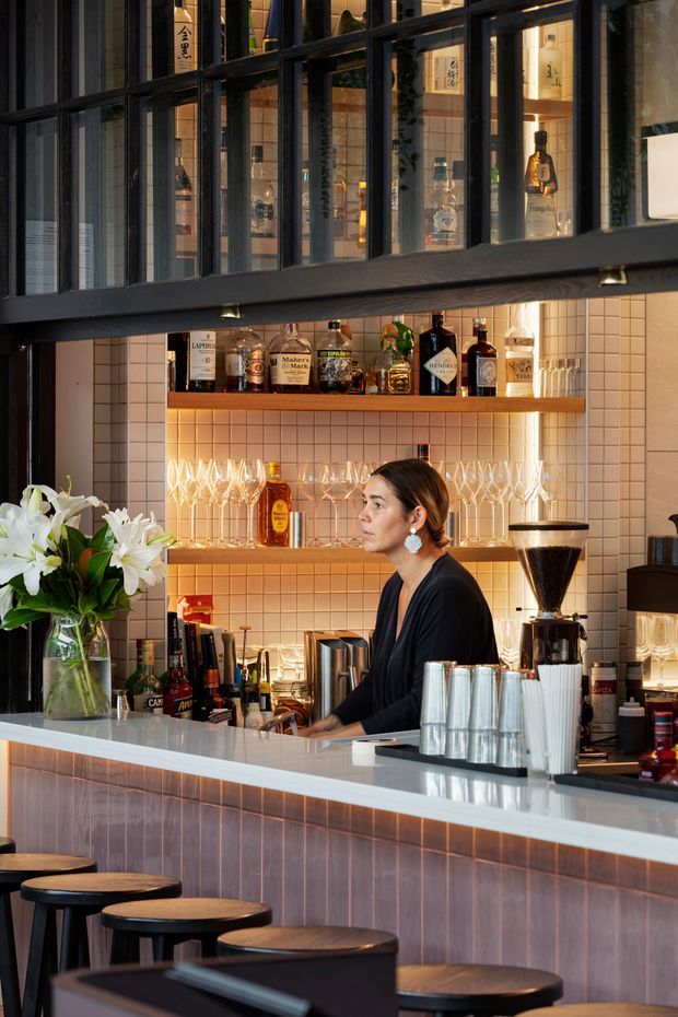 A warmly lit contemporary bar and café interior viewed from the customer side of the counter. The space features a long tiled bar front and countertop, dark-framed glazed upper partition, illuminated timber liquor shelves, pale square wall tiles, hanging glassware, a coffee grinder, bottles, and black stools with round timber seats. A bartender is working behind the counter.
