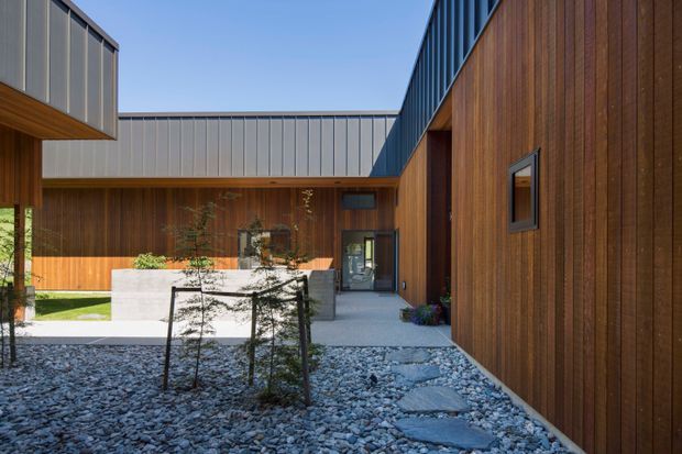 Modern courtyard exterior with dark timber-clad walls, standing-seam metal roof and fascia elements, and a gravel landscape with stepping stones. A concrete planter and low courtyard wall sit near the entrance, with strong geometric forms and clean contemporary detailing.