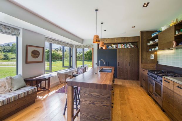 A bright open-plan kitchen and dining area with a timber island, integrated sink, dark timber cabinetry, open shelving, tiled splashback, stainless steel range cooker, and warm timber flooring. Full-height glazing opens the room to a green rural landscape, while a built-in window seat and dining nook add a relaxed residential feel. Pendant lights and recessed ceiling lights illuminate the space.