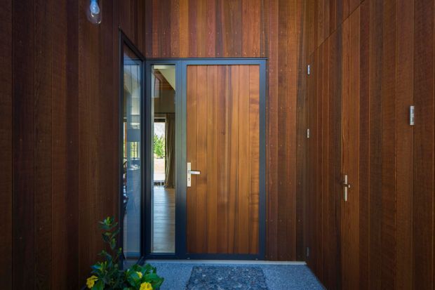 A compact entryway clad almost entirely in rich vertical timber wall panelling, with a large timber front door set within dark metal-framed glazing. The partially open glazed side panel reveals a timber-floored interior beyond. A dark textured threshold and small planted arrangement add contrast.