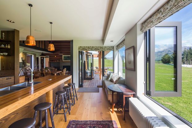A bright, elongated open-plan kitchen and living area with a substantial timber island, dark timber cabinetry, integrated shelving, pendant lighting, bar stools, and a compact seating nook beside large windows. The space opens toward a timber deck and expansive green landscape with mountain views. Warm timber flooring and joinery contrast with light walls and abundant natural light.