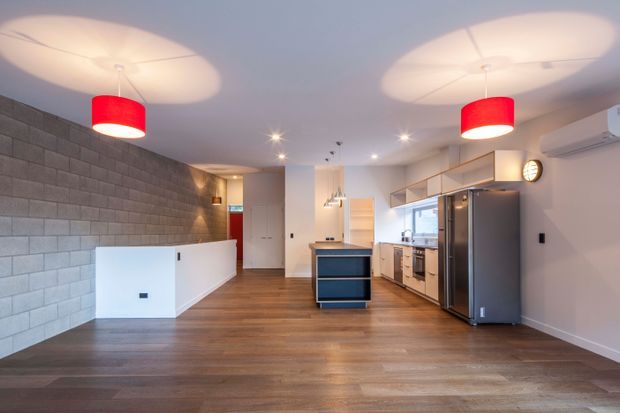 Open-plan contemporary kitchen and living space with warm timber flooring, a dark grey block feature wall, white cabinetry, stainless-steel appliances, a central island, and striking red pendant lights. The room is bright and spacious with a clean, refined fit-out.