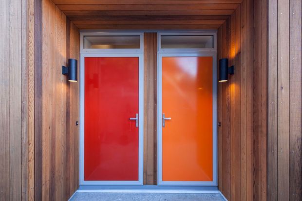 A close-up architectural view of two vivid red-orange entry doors set side by side within a recessed, richly timber-lined entrance. The doors have silver metal frames, narrow glazed transoms above, dark wall-mounted sconces, and a grey threshold floor. Vertical timber cladding and a layered timber soffit create a warm, symmetrical facade detail.