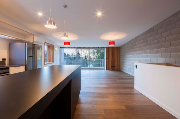 Open-plan contemporary kitchen and living area with a large dark island bench, integrated cabinetry and appliances, warm timber flooring, a textured grey masonry feature wall, pendant lighting, and full-width glazing opening to a balcony or exterior view.