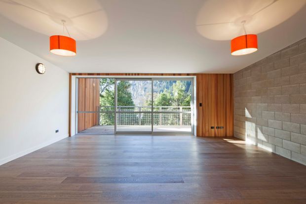 An empty, light-filled contemporary living room with wide timber flooring, a white ceiling, exposed grey concrete-block feature wall, and warm timber wall panelling around large sliding glass doors. Two bright orange pendant lights hang from the ceiling. The glazing opens to a balcony or terrace with leafy mountainous scenery beyond.