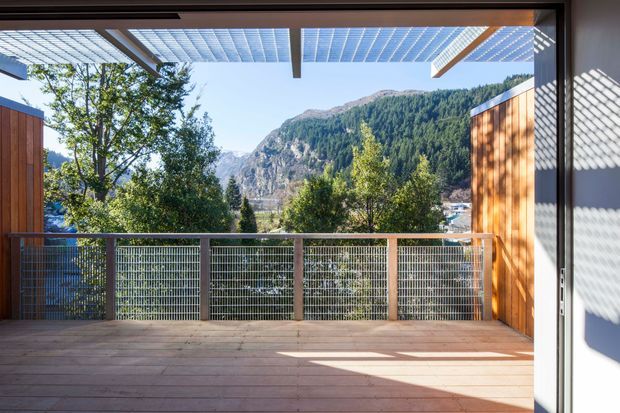 A covered timber deck or terrace viewed from inside, framed by large openings and warm vertical timber cladding. The deck has timber flooring, a metal mesh balustrade, and a translucent slatted pergola overhead. Beyond it is a dramatic forested mountain landscape, while strong sunlight creates patterned shadows across the deck and wall.