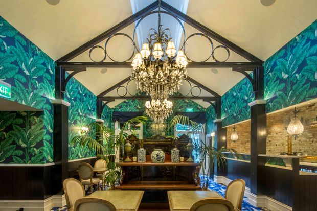 A richly decorated, symmetrical hospitality-style interior with a high vaulted ceiling, exposed dark metal trusses and circular detailing, ornate crystal chandeliers, bold tropical botanical wallpaper, dark wall panelling, exposed brick, palms, and classic furnishings. A large console and decorative mirror anchor the far end of the space, while illuminated wall niches and seating line the sides.