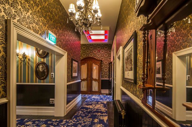 A richly decorated, narrow hallway with ornate dark floral wallpaper, dark timber wainscoting, cream moulded door surrounds, decorative mirrors, framed artwork, and a patterned blue carpet. A traditional chandelier hangs centrally, with a carved wooden armoire at the far end and an illuminated exit sign above a doorway. The atmosphere is theatrical, opulent, and strongly heritage-inspired.