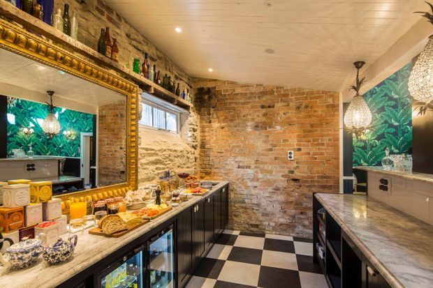 A richly styled compact home bar or entertaining space with exposed brick walls, a black-and-white checkerboard floor, dark built-in cabinetry, stone benchtops, open bottle shelving, a large ornate gold-framed mirror, patterned botanical wallpaper, and decorative pendant lighting. The space includes beverage refrigerators and extensive bar storage.