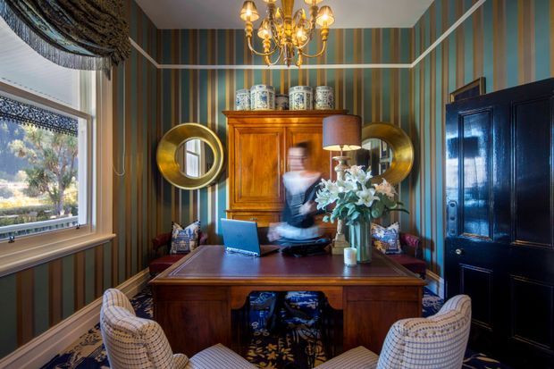 A richly decorated traditional home office with a dark wood desk, matching timber cabinetry, patterned upholstered chairs, striped teal and brown wallpaper, ornate brass chandelier, round wall mirrors, and a large window with layered drapery. The room has a warm, formal, eclectic character.