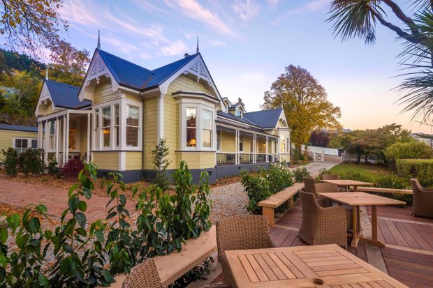 Exterior view of a restored Victorian-era villa with ornate gabled rooflines, decorative timber fretwork, bay windows, pale weatherboard cladding, and a covered verandah. The house sits within landscaped gardens and opens onto a timber outdoor dining deck with tables and chairs.