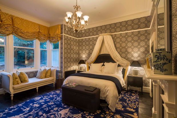 Luxurious traditional bedroom with a four-poster-style bed dressed in draped fabric, ornate patterned wallpaper, a deep blue floral carpet, upholstered window seat, heavy gold-toned Roman blinds, decorative chandelier, and detailed white trim and built-in cabinetry.