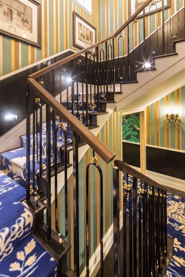 A richly decorated multi-level stairway with dark timber handrails, glossy black balusters, and brass finials. The stairs are covered in a bold navy and cream patterned carpet, while the walls feature vertical green, cream, and ochre striped wallpaper with dark wainscoting. Framed artwork, decorative lighting, and a vivid botanical artwork add a highly ornate, theatrical character.