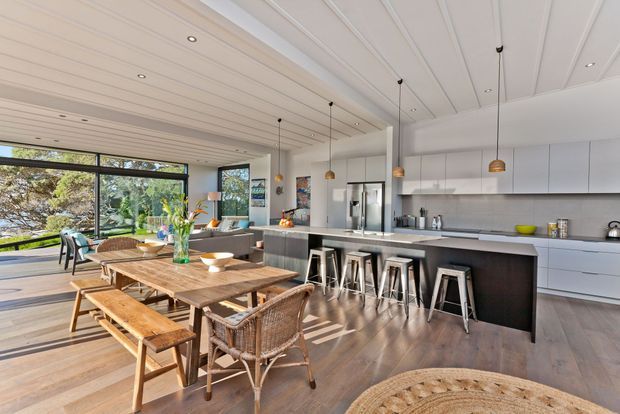 Bright open-plan kitchen and dining area with a large dark island, white full-height cabinetry, integrated appliances, pendant lights, timber flooring, and a long rustic timber dining table with bench seating and woven chairs. Expansive glazing opens to a leafy outdoor setting, while the white panelled ceiling adds architectural character.