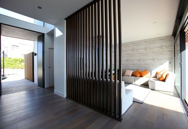 Bright contemporary home interior showing an entryway with dark timber flooring and a full-height dark timber vertical-slat screen dividing it from a sunlit living room. The living area contains a light sectional sofa with orange and neutral cushions, extensive glazing, and a partially visible timber-clad wall.