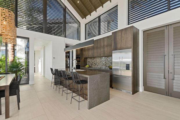 A bright, open-plan contemporary kitchen with a large stone island and seating for four. Dark timber-look cabinetry, a textured stone backsplash, stainless-steel appliances, and extensive clerestory-style louvred glazing sit beneath a high pitched timber ceiling. A dining area and planted courtyard-like space are visible at the left.