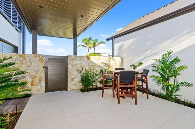 A bright, modern outdoor courtyard or patio dining area enclosed by rendered walls and a natural stone feature wall. The space has large light-coloured tiled paving, a timber dining table and chairs, tropical planting, a dark metal door, and a timber-lined covered ceiling with recessed lighting. A small pool or water feature is partially visible at the left edge.