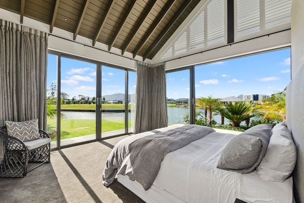 Bright, refined bedroom with a large bed positioned beside expansive corner glazing overlooking a landscaped waterfront setting. The room features a dramatic pitched ceiling with exposed timber rafters, dark timber ceiling boards, full-height grey curtains, plantation shutters, and neutral layered bedding. An accent chair sits in the corner, while strong natural light creates a calm, airy atmosphere.