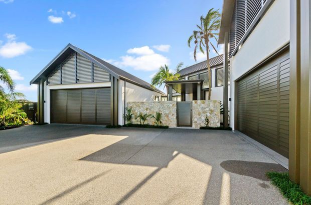 Bright exterior view of a contemporary multi-building residential complex, showing several attached garages with dark horizontal doors, steep gabled roofs, louvred upper facade panels, pale rendered walls, and a low irregular-stone privacy wall around a pedestrian entry. A broad concrete-paved driveway or courtyard occupies the foreground, with tropical palms visible around the property.