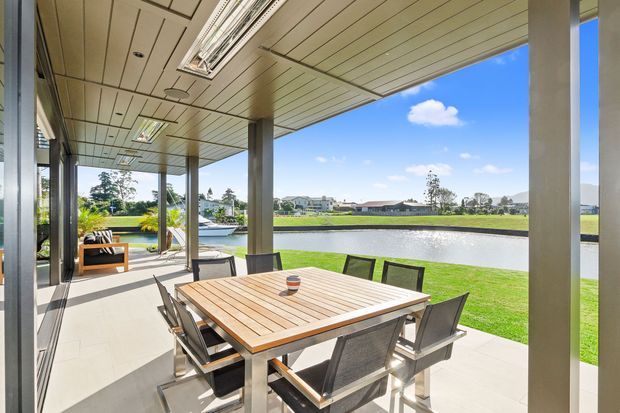 Bright covered outdoor dining terrace overlooking a landscaped lake or waterway. The space has a large timber dining table with contemporary outdoor chairs, a tiled floor, slim structural columns, extensive glazing, and a linear panelled ceiling with recessed lighting. The setting is open, airy, and modern, with lawn and low-rise residential buildings visible beyond the water.