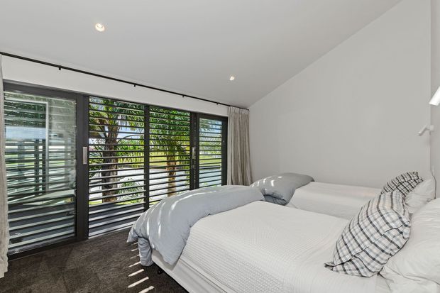 Bright contemporary bedroom with two beds dressed in white bedding, grey throws, and checked accent cushions. Full-height dark-framed louvred glazing provides filtered natural light and views of greenery, while recessed ceiling lights and neutral finishes create a clean, relaxed atmosphere.