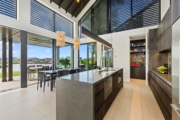 A bright, contemporary open-plan kitchen with a large stone island, dark timber cabinetry, integrated appliances, and pendant lights. Floor-to-ceiling glazing and high clerestory windows connect the kitchen and dining area to an outdoor terrace, while exposed dark ceiling structure and horizontal blinds add architectural definition.