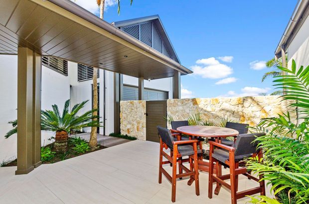 A bright modern courtyard or patio entertaining area beside a contemporary residence. The space features a covered outdoor dining zone with a timber high table and chairs, pale tiled or concrete-look paving, a stone feature wall, louvred screening, and lush tropical planting. Strong sunlight and clear blue sky create an airy resort-like atmosphere.
