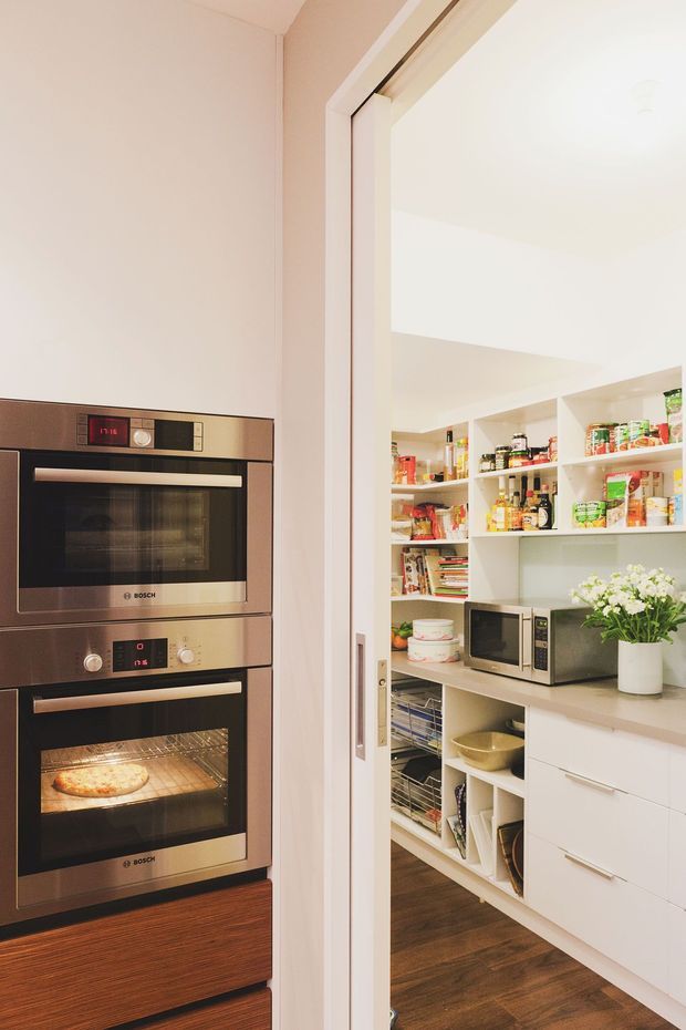 View from a kitchen through a wide doorway into a well-organised walk-in pantry. The foreground contains stacked built-in Bosch ovens with dark wood cabinetry, while the pantry has white open shelving and lower drawers stocked with food, a microwave, countertop appliances, and a timber-look floor. The space is bright, warm, and highly functional.
