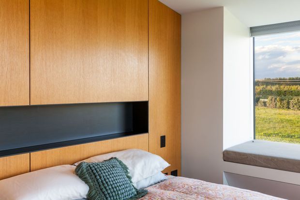 A contemporary bedroom with extensive warm timber built-in cabinetry forming a headboard wall, including a dark recessed display niche and integrated black switches. The bed is partially visible with white bedding, a textured teal cushion, and a muted patterned throw. A large corner window provides views over a green rural landscape, while a built-in window seat sits beneath it.