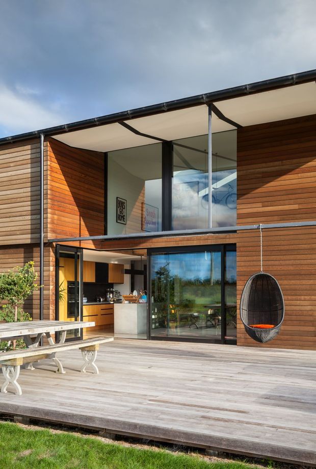 A modern two-storey timber-clad home opening onto a broad timber deck. The facade features horizontal wood cladding, large black-framed glazed doors and windows, an upper-level glass-fronted space, and a covered terrace roof. An outdoor dining table and hanging egg chair occupy the deck, while a compact kitchen is visible through the open glazing.