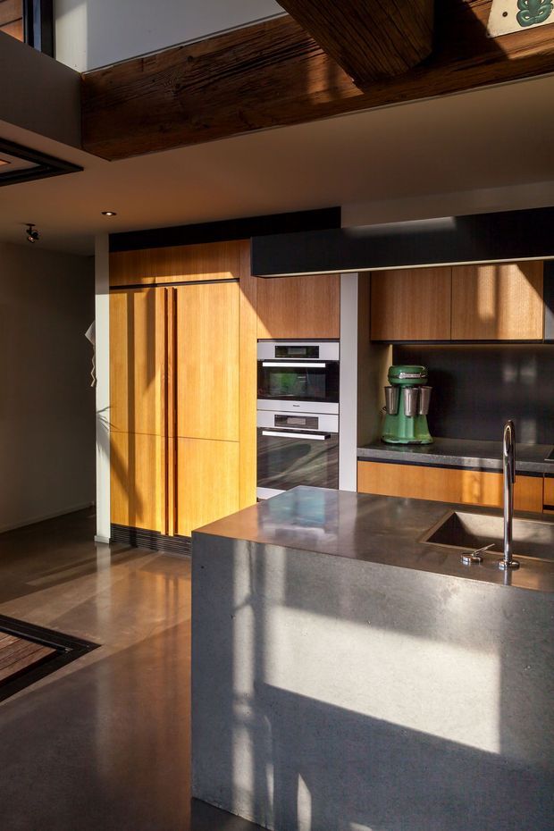 A modern kitchen with warm timber cabinetry, integrated double ovens, dark benchtops and splashback, and a large concrete island with an inset sink and tall chrome tap. Strong sunlight creates dramatic shadows across the cabinetry and island, while exposed dark timber ceiling beams add warmth and character.