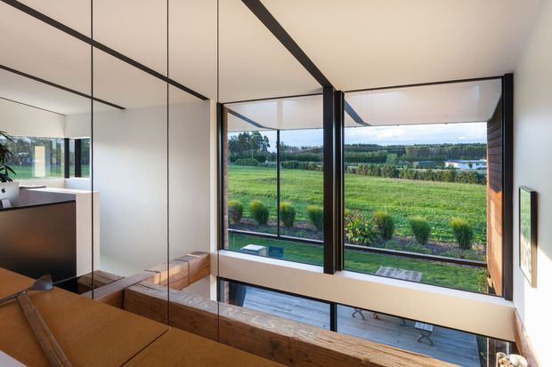 A contemporary interior viewed from a dining or living area, featuring full-height black-framed glazing overlooking open green countryside and water beyond. Warm timber flooring and a timber dining table contrast with white walls, black structural lines, and built-in cabinetry. A large mirror reflects the room and reinforces the sense of openness and connection to the landscape.