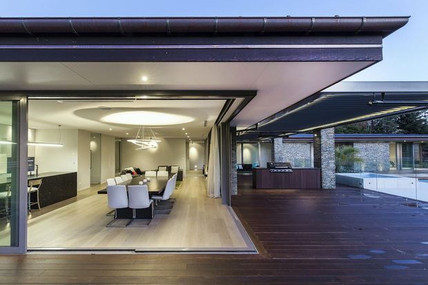Contemporary single-level residential entertaining area with a large open-plan dining and kitchen interior opening directly onto a broad timber deck. The design features extensive retractable glazing, a deep roof overhang, dark timber decking, stone-clad columns, integrated outdoor kitchen cabinetry, and a swimming pool visible to the right. Inside, a long dining table, upholstered dining chairs, pale timber flooring, recessed lighting, and a sculptural ceiling light create a refined indoor-outdoor connection.