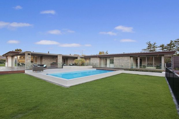 Single-storey low-slung residence viewed from the landscaped rear yard, with a broad flat roof, extensive glazing, masonry walls, covered outdoor living area, and a rectangular swimming pool set into a paved terrace. The architecture has a clean mid-century-influenced modern character, with generous lawn and minimal planting.