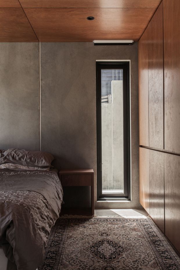 A compact, moody bedroom with a partially visible bed on the left, dark textured bedding, a small built-in bedside table, and full-height timber wardrobe cabinetry along the right wall. The room has dark grey concrete-like wall panels, warm timber ceiling and joinery, a narrow vertical window with a dark frame, and a patterned Persian-style rug. Natural light enters through the window, creating a warm, intimate atmosphere.