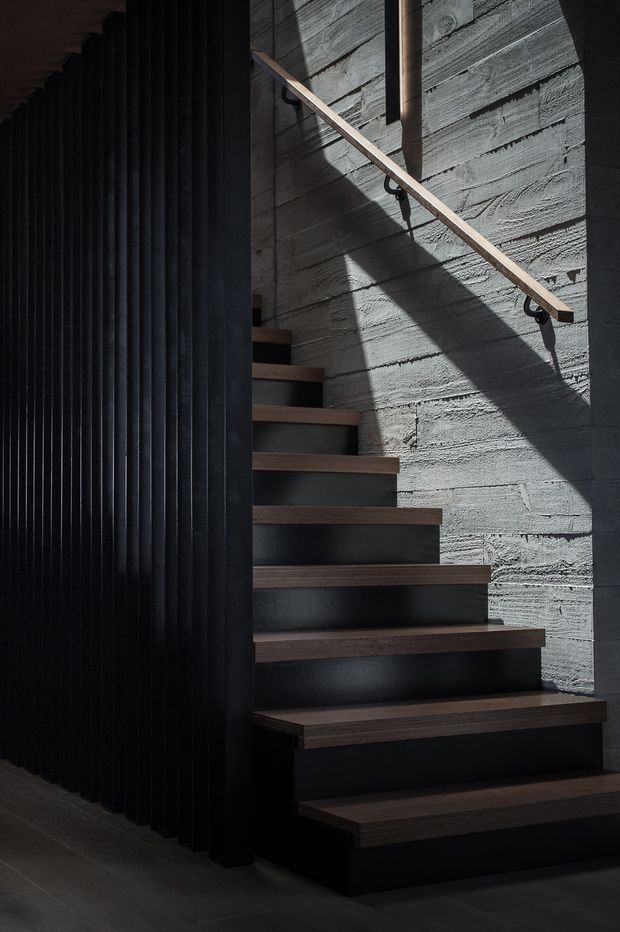 A dramatic contemporary interior stairway with floating dark timber treads set against a textured grey concrete wall. A simple timber handrail follows the stair, while a tall black vertical slatted screen forms the left side. Strong directional light creates geometric shadows across the wall, emphasizing the moody, sculptural material palette.