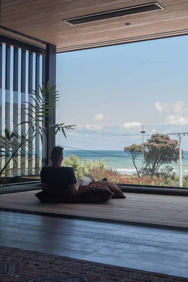 A calm, light-filled indoor lounge or sunroom opens directly onto a broad ocean view through floor-to-ceiling glazing. The space features dark timber flooring, a timber-lined ceiling with a recessed linear ventilation grille, dark structural framing, clear glass balustrade or glazing, and a low floor-level seating arrangement with cushions. A tall indoor plant sits beside the glazing, while coastal vegetation, beach, and ocean are visible outside.