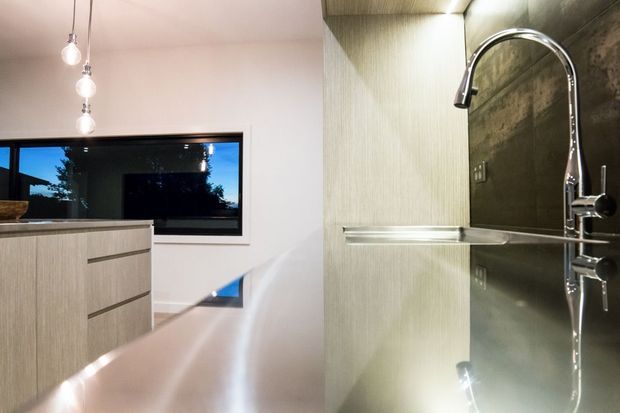 Contemporary kitchen interior with light timber-grain cabinetry, a dark reflective stone benchtop and matching dark splashback, chrome gooseneck tap, and a minimalist cluster of pendant lights. A large black-framed window provides a view outside, while integrated under-cabinet lighting highlights the work surface.