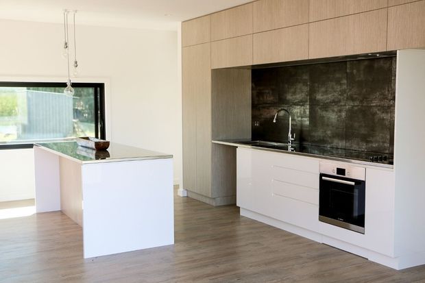 Bright contemporary kitchen with a large white island, pale timber full-height cabinetry, white lower cabinets, a dark textured splashback, integrated oven and cooktop, and timber-look flooring. A wide window and pendant lights add natural light to the open space.