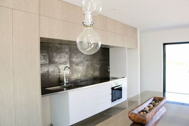 Bright contemporary kitchen with pale timber-look overhead cabinetry, white lower cabinetry, a dark reflective splashback, integrated oven and cooktop, stainless steel sink and tap, and a long stone-look island or peninsula. Clear glass pendant lights hang above the island, with large glazing or a glass door at the right.