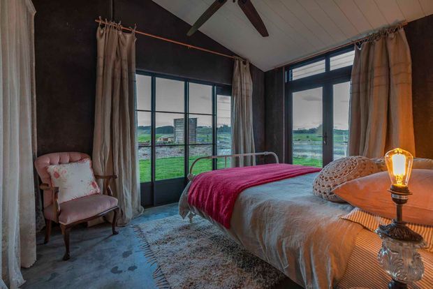 A warm, rustic bedroom with a metal-framed bed, layered neutral bedding and a deep red throw. Full-height dark-framed glazing and glass doors open the room to expansive green rural views. Heavy linen curtains, a vintage upholstered armchair, exposed dark wall surfaces, timber ceiling boards and an illuminated decorative bedside lamp create a cozy farmhouse-like atmosphere.