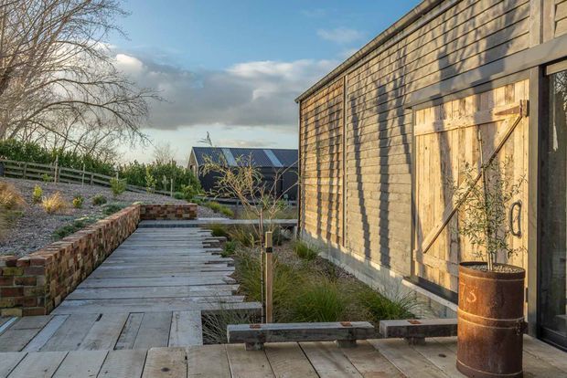 A rustic landscaped outdoor walkway beside a timber-clad building, with weathered timber decking and steps, brick retaining walls, native-style planting, and a large barn-style timber door. The scene has a warm, textural rural character under clear evening light.